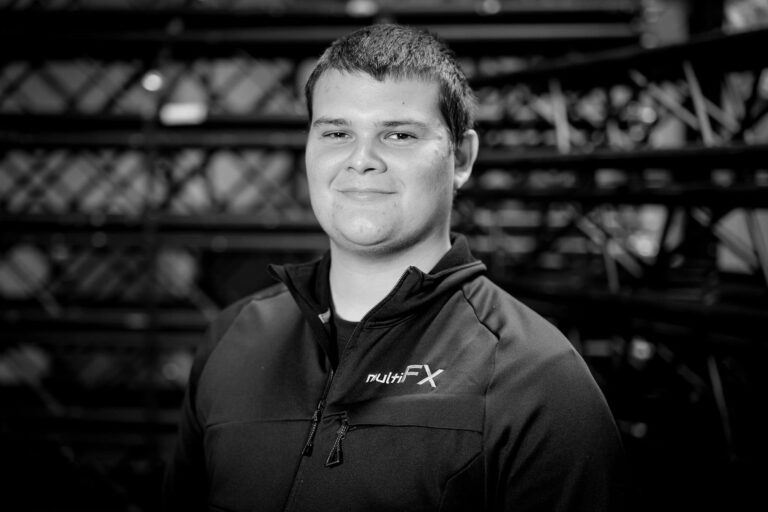 A young person with short dark hair smiles at the camera, wearing a zip-up jacket with the “nutriFX” logo, standing in front of a blurred background with metal structures. The image is in black and white.