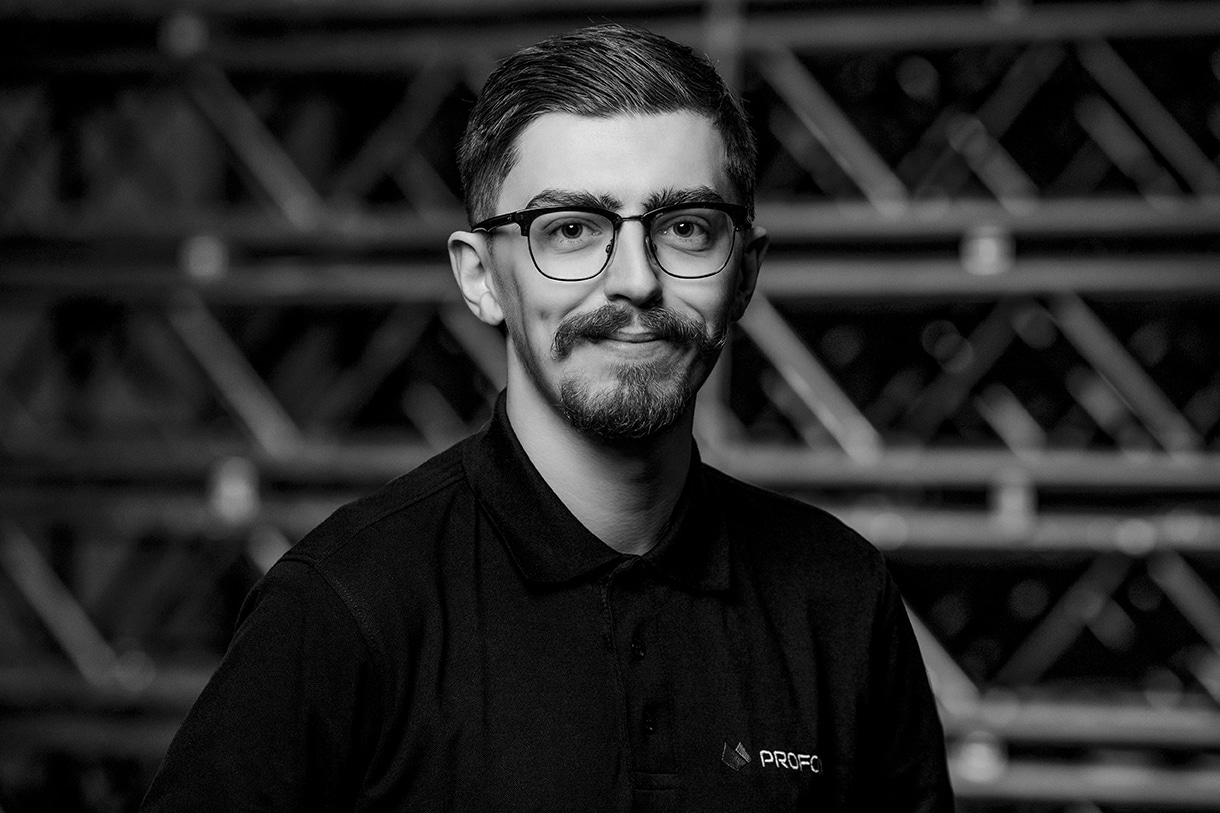 A young man with glasses, short hair, and a trimmed beard is wearing a dark collared shirt and is smiling slightly while looking at the camera. The background is blurred with a pattern of intersecting lines.
