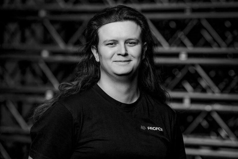 A person with long hair, wearing a dark FROG FX t-shirt, smiles while standing in front of a background featuring diagonal metal bars. The image is in black and white.