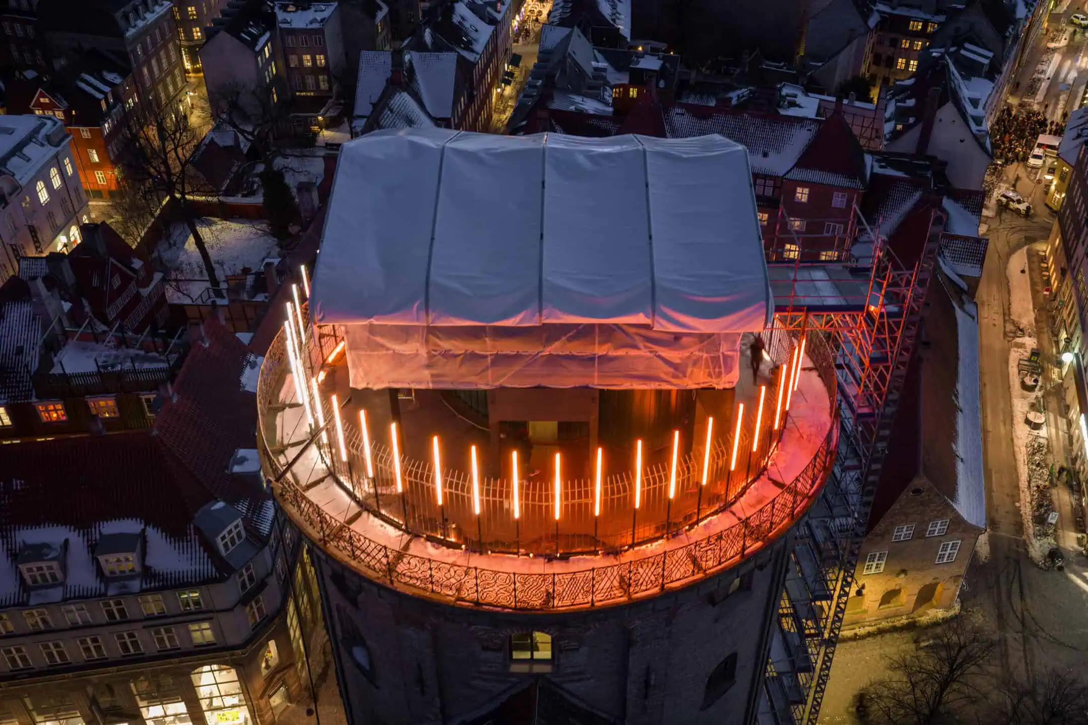 Aerial view of a round tower with orange lights and a white-covered structure on top, surrounded by snowy rooftops and city streets at dusk.