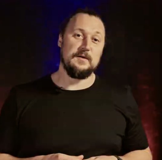 A man with a beard and hair pulled back in a black t-shirt standing in front of a blue and red background.