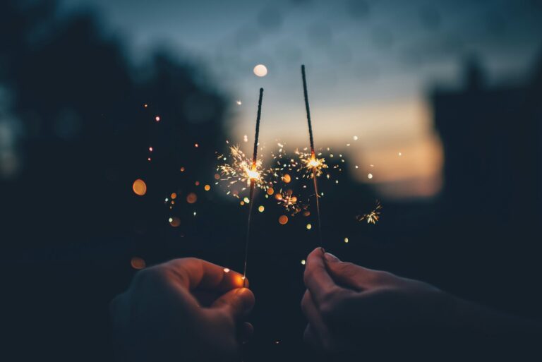 Two hands each hold a lit sparkler, with glowing sparks flying in the evening darkness and blurred lights in the background.