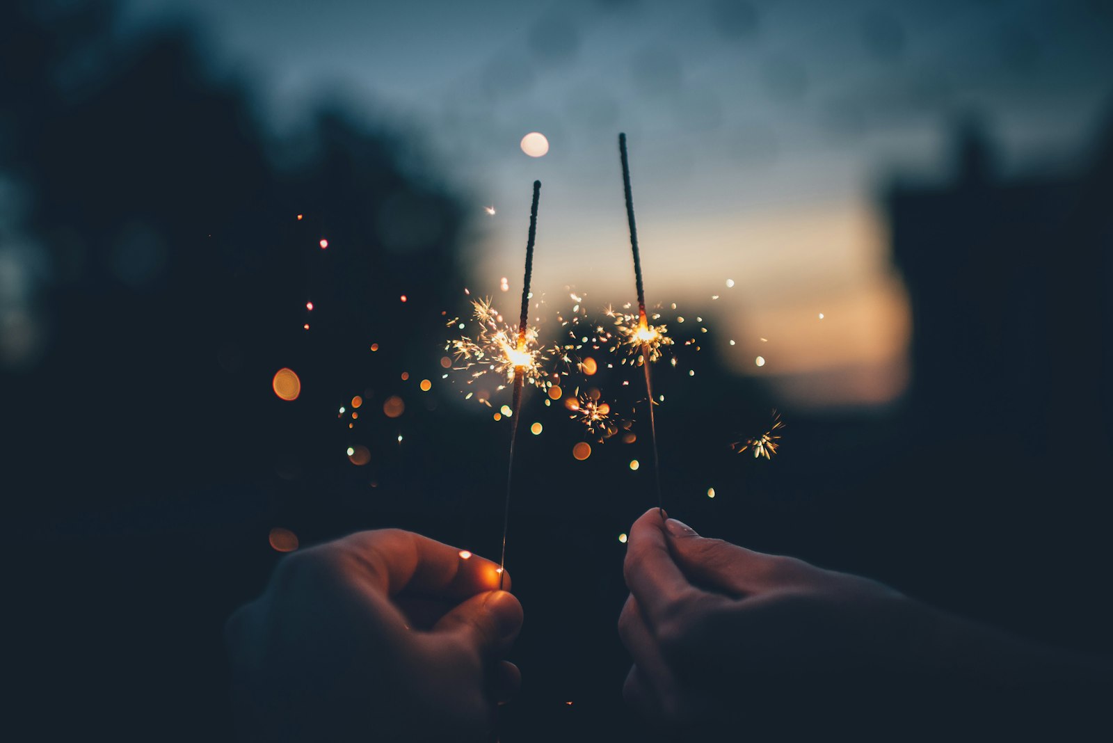 Two hands each hold a lit sparkler, with glowing sparks flying in the evening darkness and blurred lights in the background.