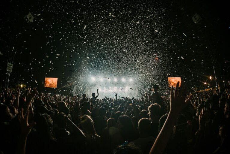 A large crowd at a nighttime concert cheers in front of a brightly lit stage, with confetti falling from above and hands raised in the air. Two video screens display the performance.