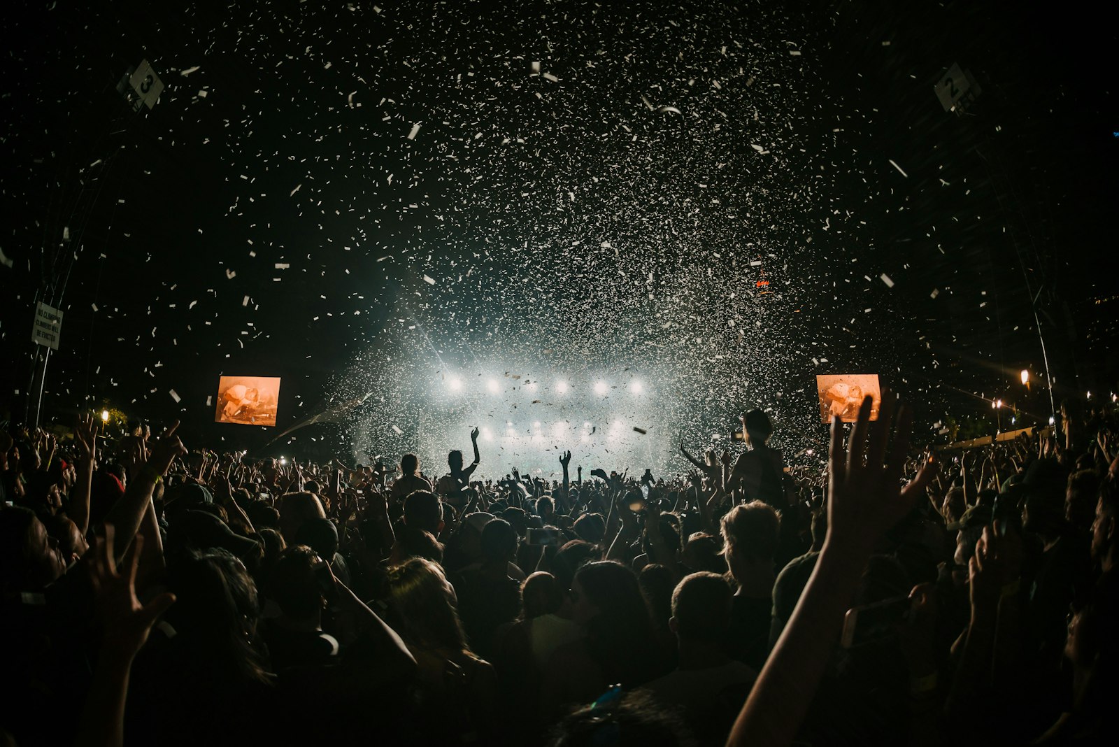 A large crowd at a nighttime concert cheers in front of a brightly lit stage, with confetti falling from above and hands raised in the air. Two video screens display the performance.