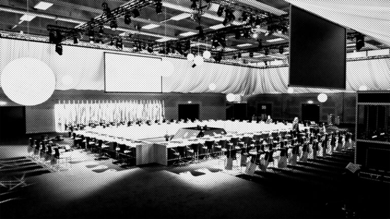 Large conference room with a square arrangement of tables and many chairs, set under bright lights and large screens. The space appears ready for a formal meeting or event, but is currently empty.