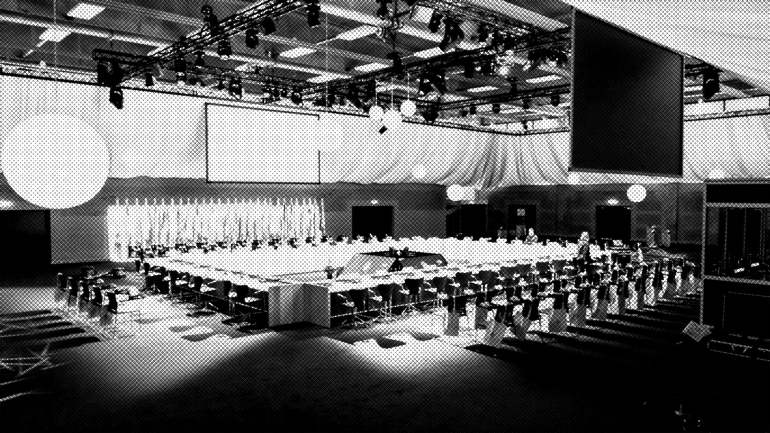 Large conference room with a square arrangement of tables and many chairs, set under bright lights and large screens. The space appears ready for a formal meeting or event, but is currently empty.