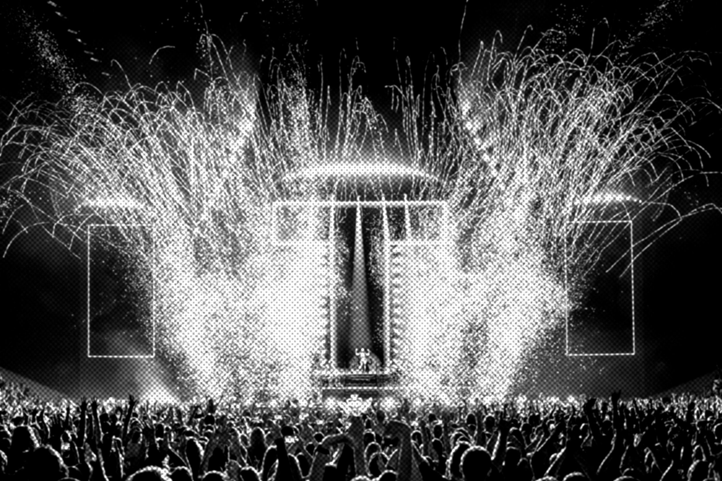 A large crowd at a concert cheers in front of a brightly lit stage with fireworks and pyrotechnics bursting in the background.