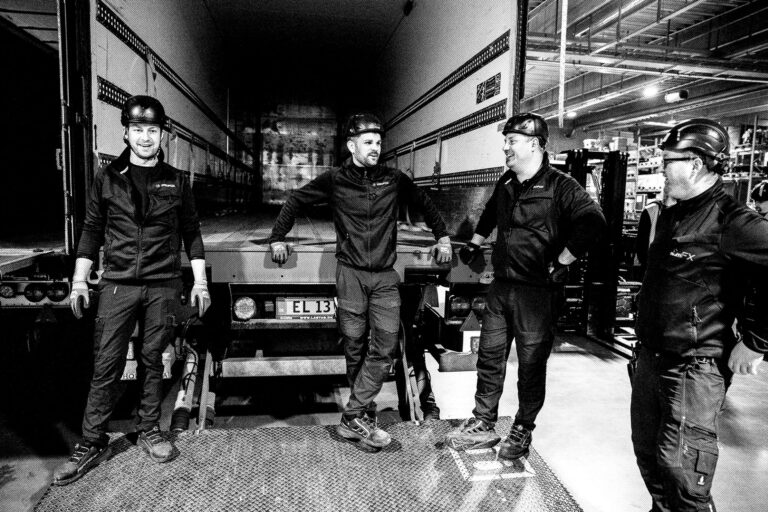 Four workers in helmets and uniforms stand and talk near the back of a truck inside a warehouse. The image is in black and white, and they appear to be taking a break together.