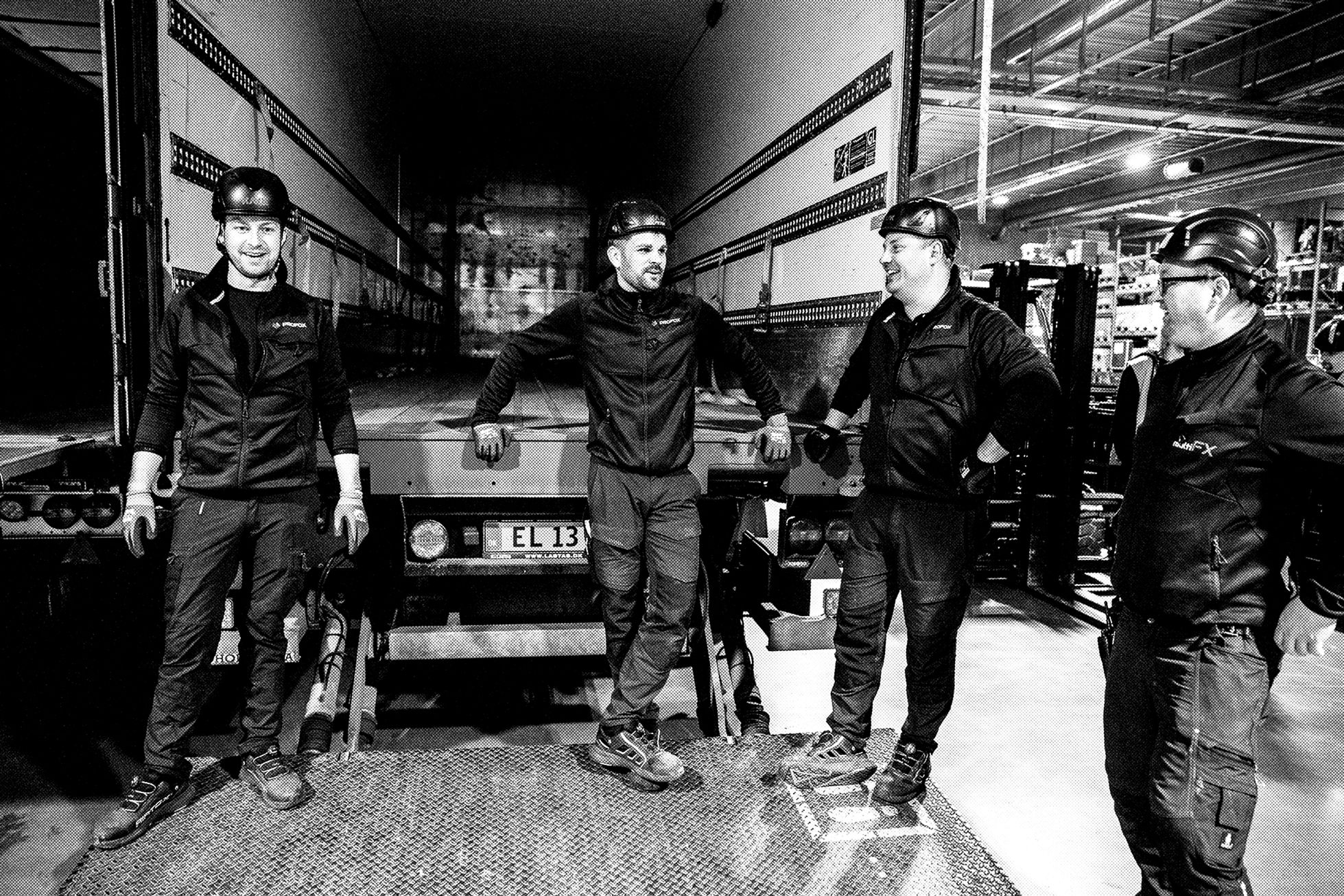 Four workers in helmets and uniforms stand and talk near the back of a truck inside a warehouse. The image is in black and white, and they appear to be taking a break together.