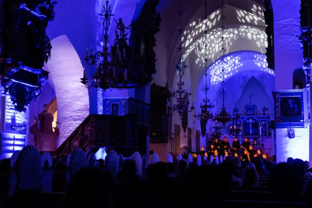An audience sits in a dimly lit church with blue and purple lighting, watching a choir perform near the altar under decorative light patterns on the arched ceiling.