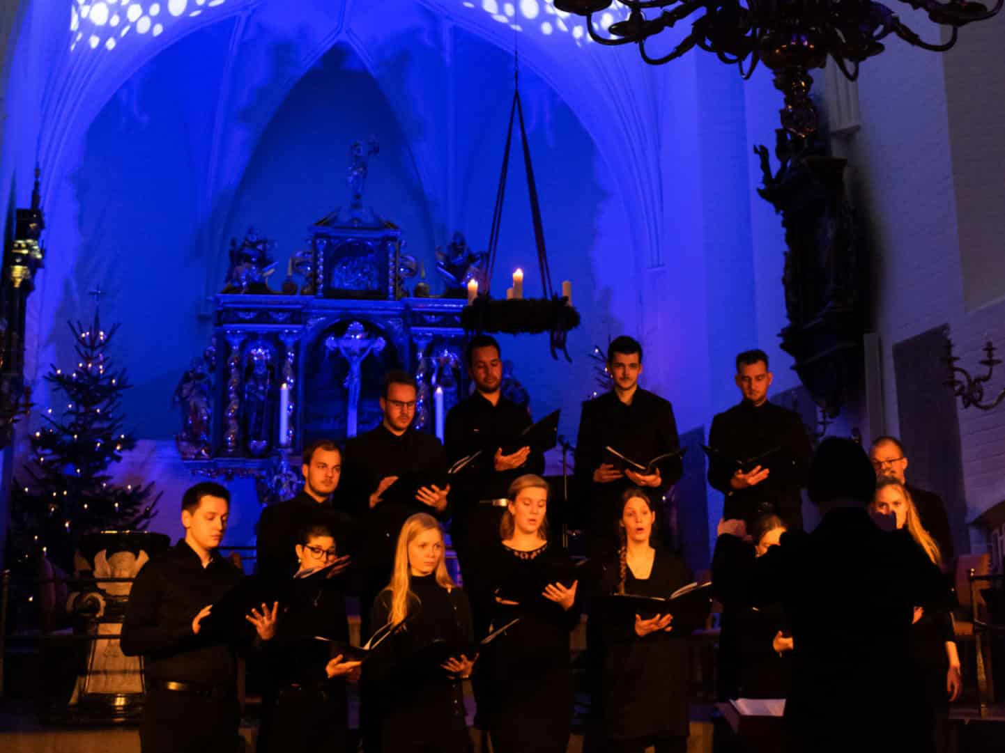 A choir dressed in black sings in a dimly lit church with blue and orange lighting, a Christmas tree, and a decorated altar in the background.