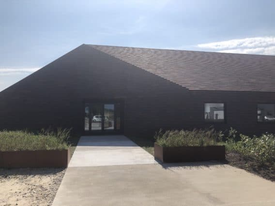 A modern, dark-brick building with a sloped roof, large entrance door, and a concrete walkway flanked by raised planters with grasses, under a partly cloudy sky.