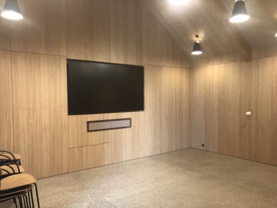 A modern, empty room with wood-paneled walls, a large flat-screen TV, ceiling lights, and a few stacked chairs in the corner.