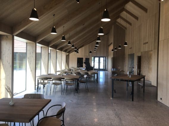 Modern dining area with wooden walls and ceiling, large windows on the left, several tables and chairs, pendant lights hanging from the ceiling, and a person standing near the far end.