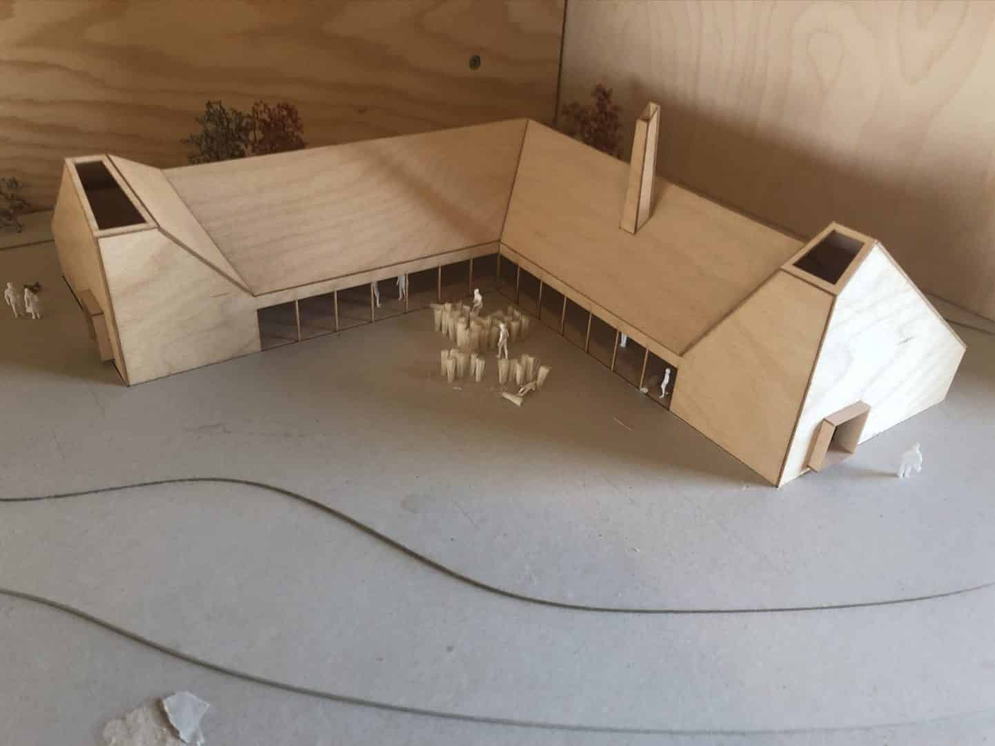 A wooden architectural model of an L-shaped building with a central courtyard, small human figures, and minimal trees, placed on a plain surface.