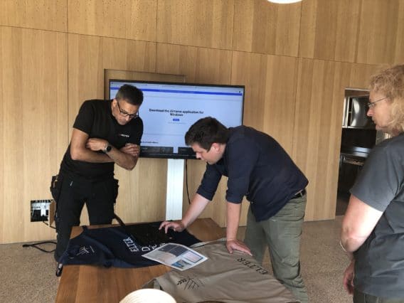 Three people stand around a wooden table with shirts and papers, looking at items. Behind them, a large screen displays a form on a website. The setting appears to be an office or meeting room with wood-paneled walls.