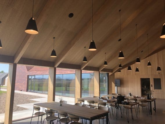 Modern dining area with wooden walls and high, sloped ceiling. Multiple pendant lights hang above tables and chairs. Large windows offer a view of grass and a nearby building, allowing natural light to fill the space.