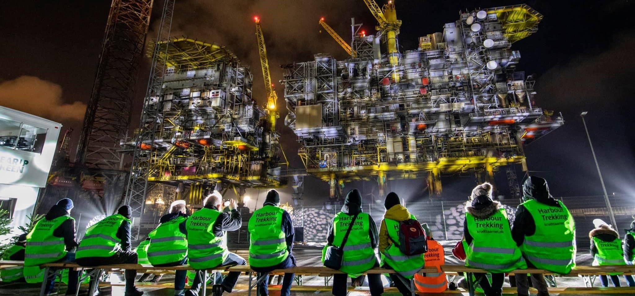A group of people in bright green safety vests sit on a bench at night, facing a large, illuminated industrial structure with cranes and scaffolding.