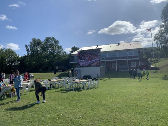 People gather on a sunny lawn with white chairs facing a large outdoor screen. A red building is in the background, and a few people are setting up or talking. Trees and a flagpole are visible.