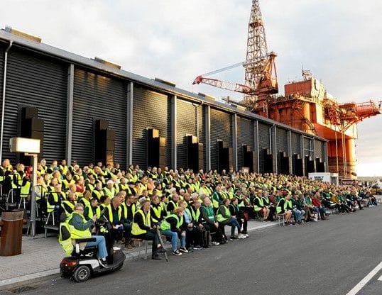 A large group of people in yellow safety vests sit outside in rows near an industrial site with a red oil rig and metal building in the background.