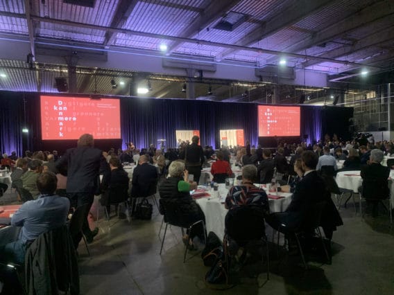 Large conference room with round tables, many seated attendees facing a stage with red screens displaying text, and a high industrial ceiling with exposed beams and lighting fixtures.