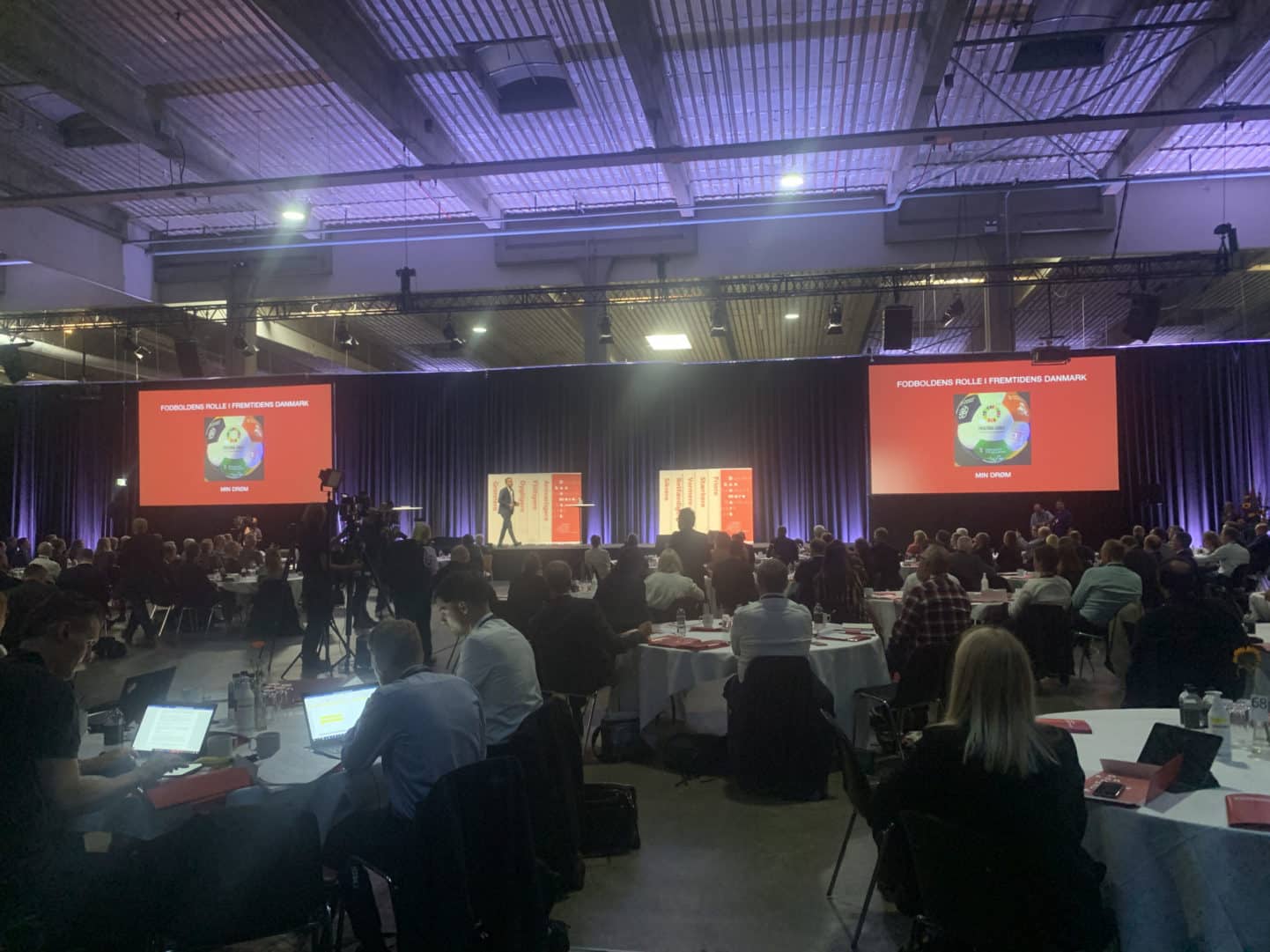 A large conference room with people seated at round tables, watching a speaker on stage. Two screens display a presentation with red backgrounds and graphics. Laptops and notebooks are visible on the tables.