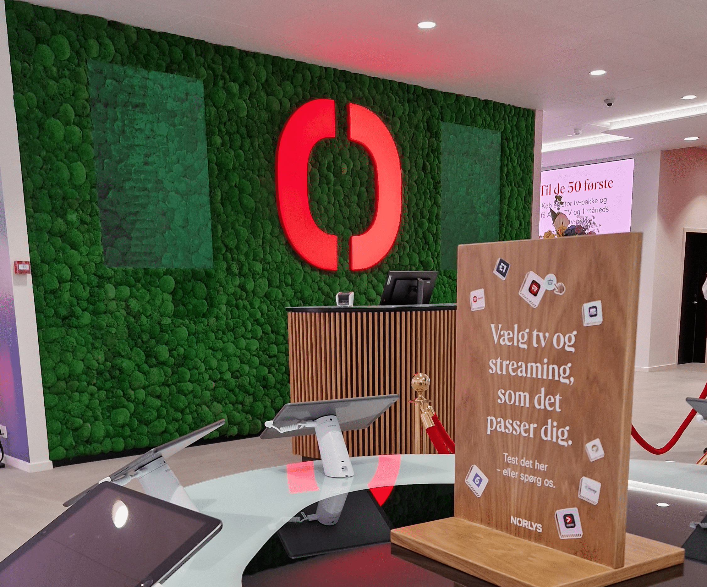 A modern store interior with a green moss wall, a large red logo, a wooden counter, and a sign in Danish offering TV and streaming options. Tablets and digital devices are on display.