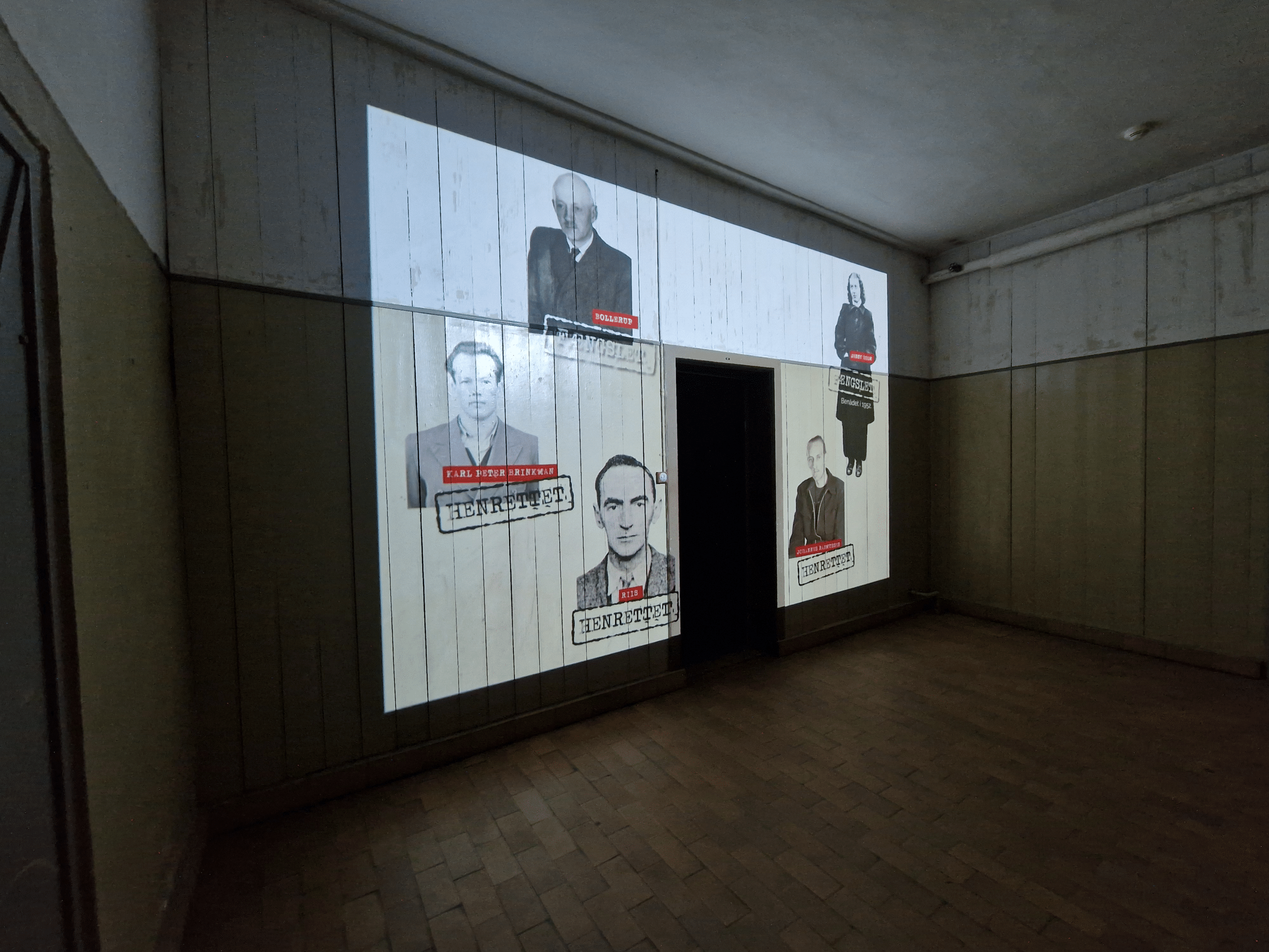 A dim room with a projection on the wall showing portraits of four people, each labeled in red and white, with a doorway in the center splitting the projection.