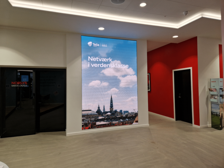 A large digital sign in a modern office displays a city skyline and Danish text: Netværk i verdensklasse. The wall behind is red, and doors and a hallway are visible beside the sign.