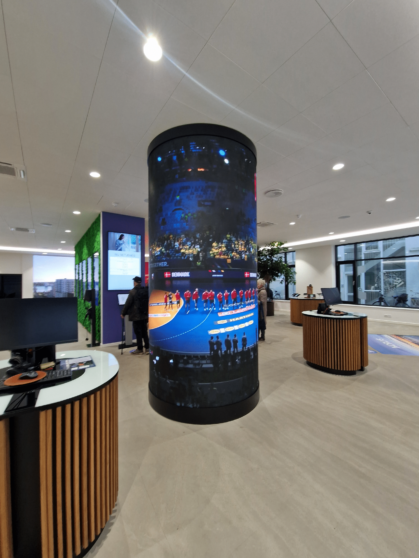 A cylindrical digital display shows a sports event in a modern, open office with desks, computers, and people interacting in the background.