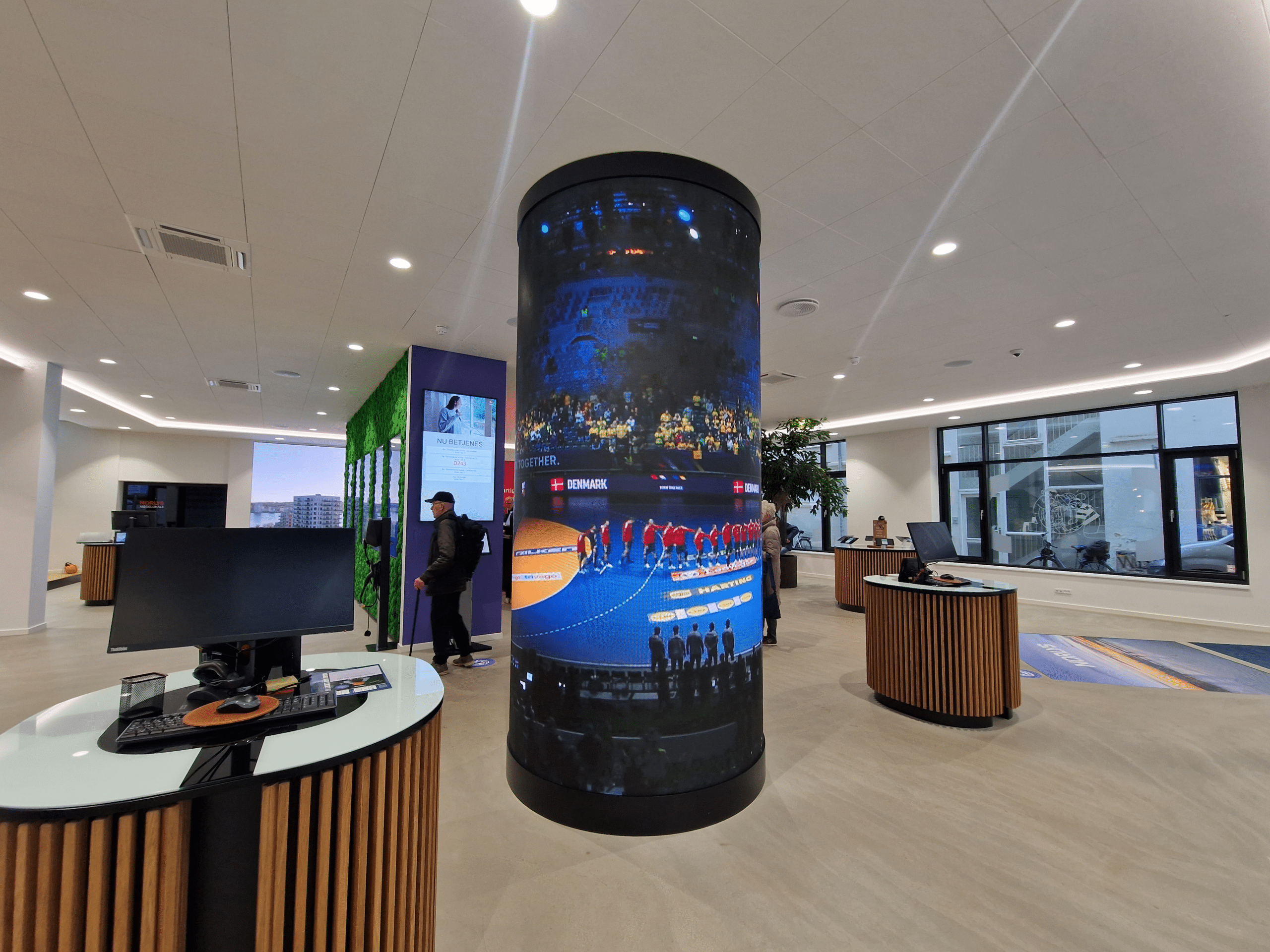 Modern office space with curved desks, large cylindrical digital display showing a sports event, one person standing near the entrance, and large windows letting in natural light.