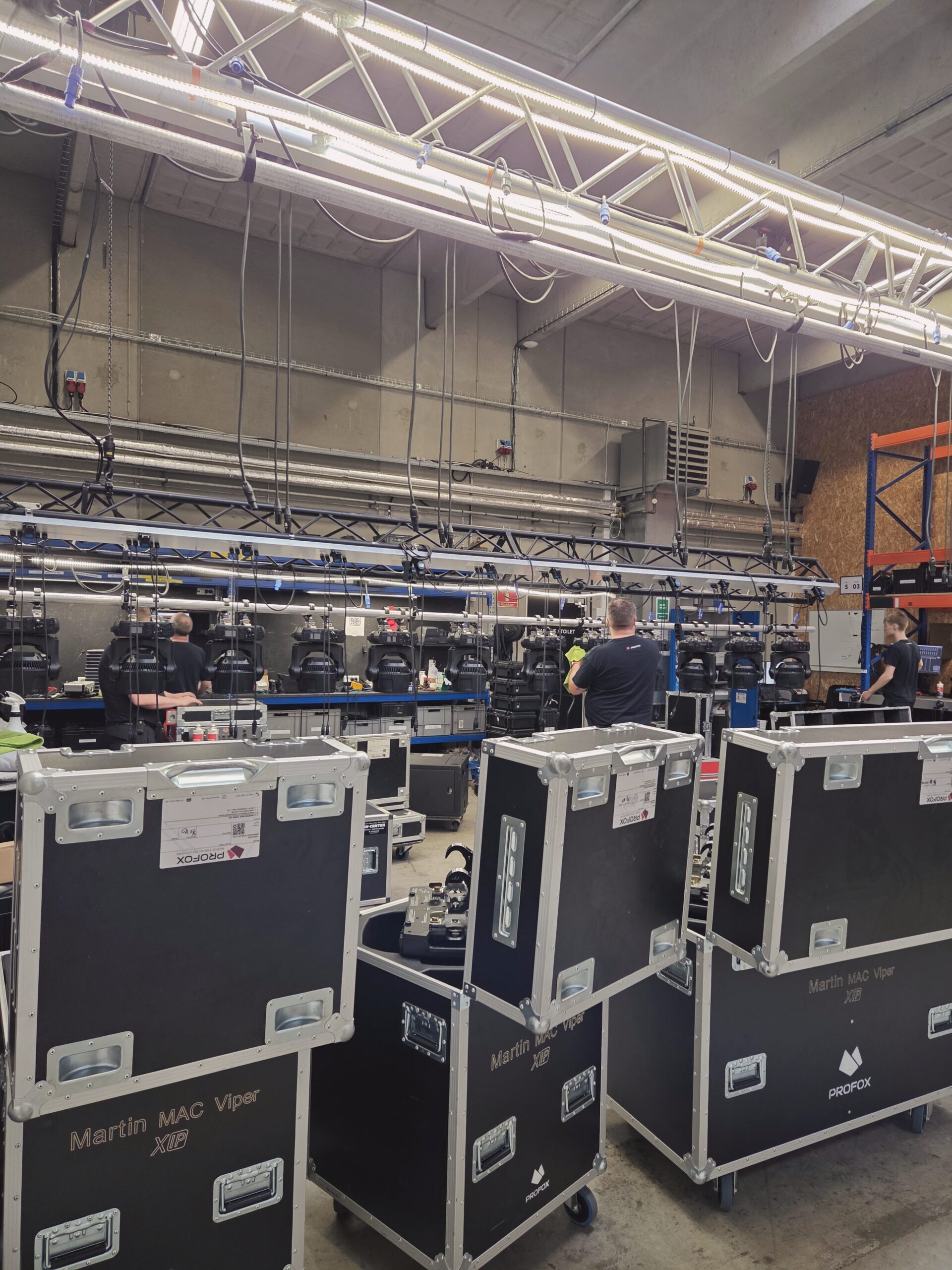 People work in a warehouse with stage lighting equipment, large black cases labeled Martin MAC Viper, cables, trusses, and shelves. The setting appears industrial, with concrete walls and various tools visible.