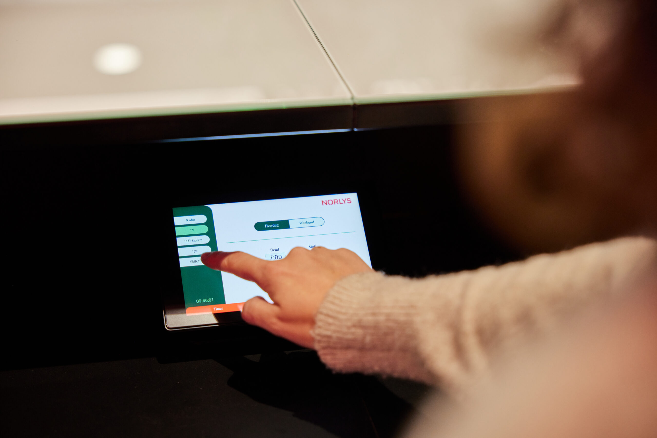 A person uses their finger to interact with a touchscreen device displaying menu options and the word NORLIS. The screen interface appears to be part of a modern digital system.