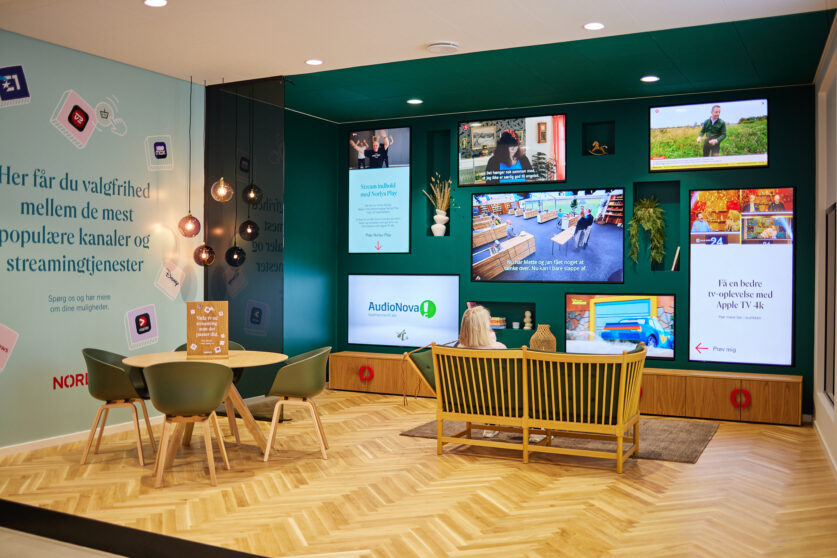 A modern lounge with green walls, a wooden floor, a round table with chairs, and a sofa facing multiple large screens displaying various shows and information. Pendant lights hang from the ceiling.