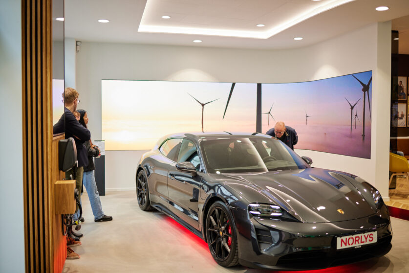 A sleek black Porsche is displayed indoors with people observing it. The background features large scenic photos of wind turbines at sunset.