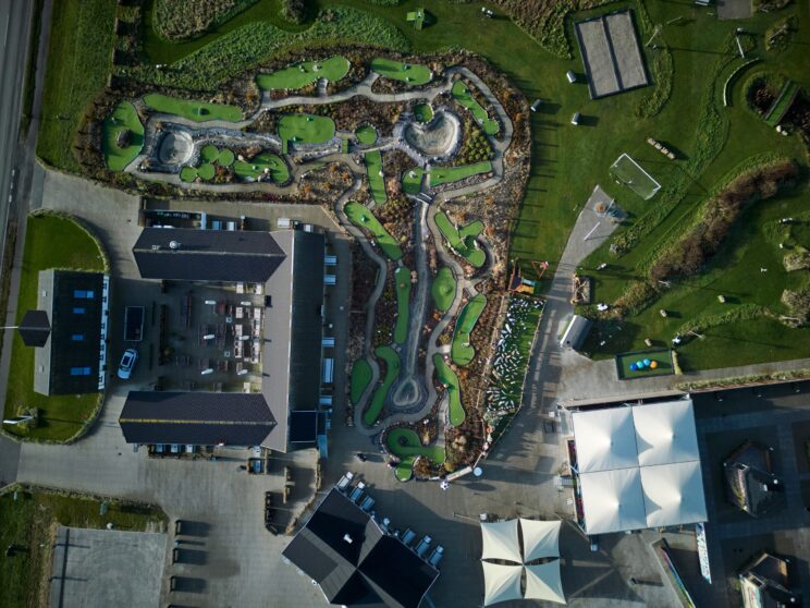 Aerial view of a mini-golf course with winding paths, green turf, rocks, and adjacent buildings, parking area, and covered outdoor seating. Grass and a road are visible on the left side.