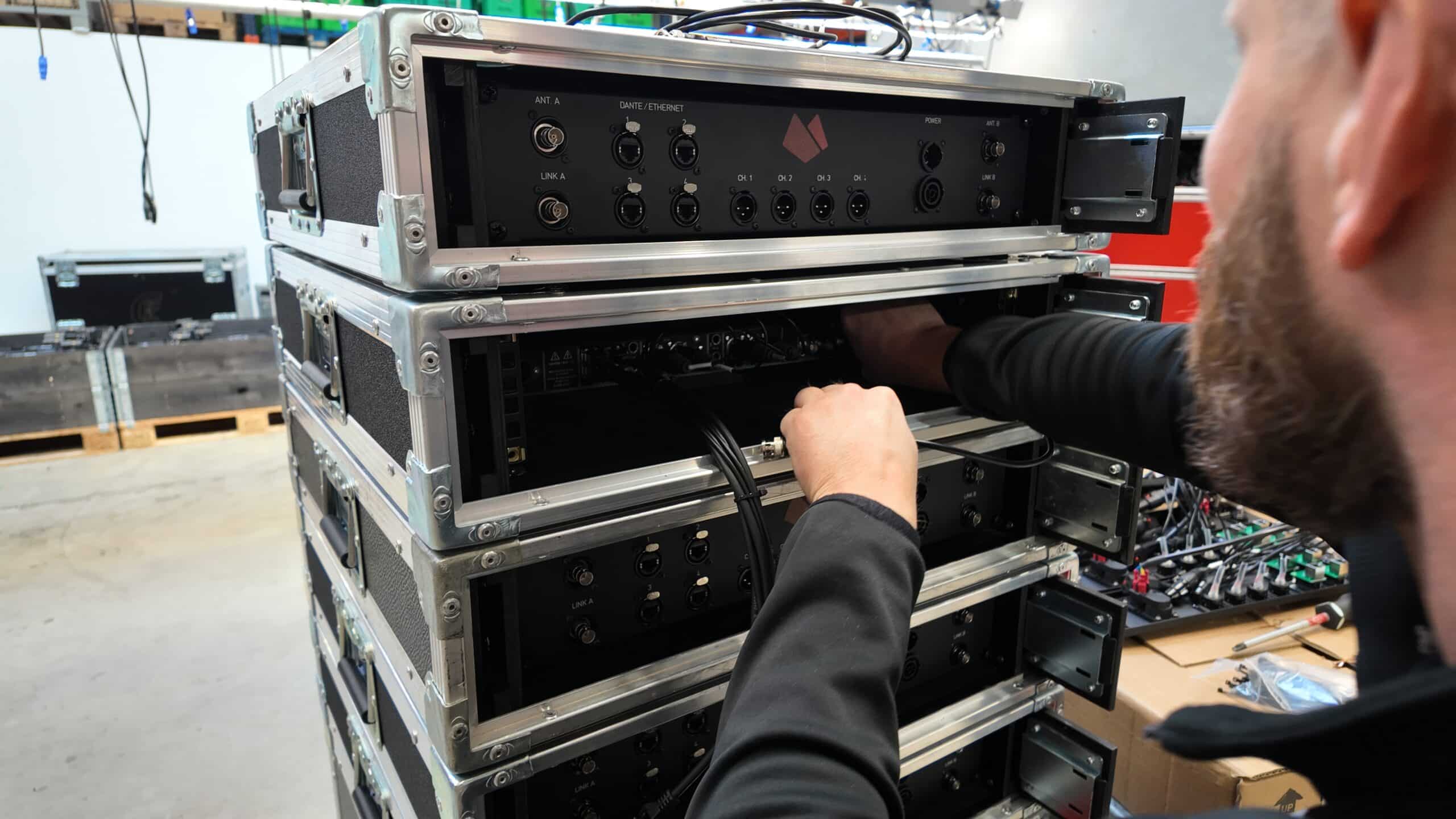 A person connects cables to the back of stacked audio equipment racks in an industrial setting.