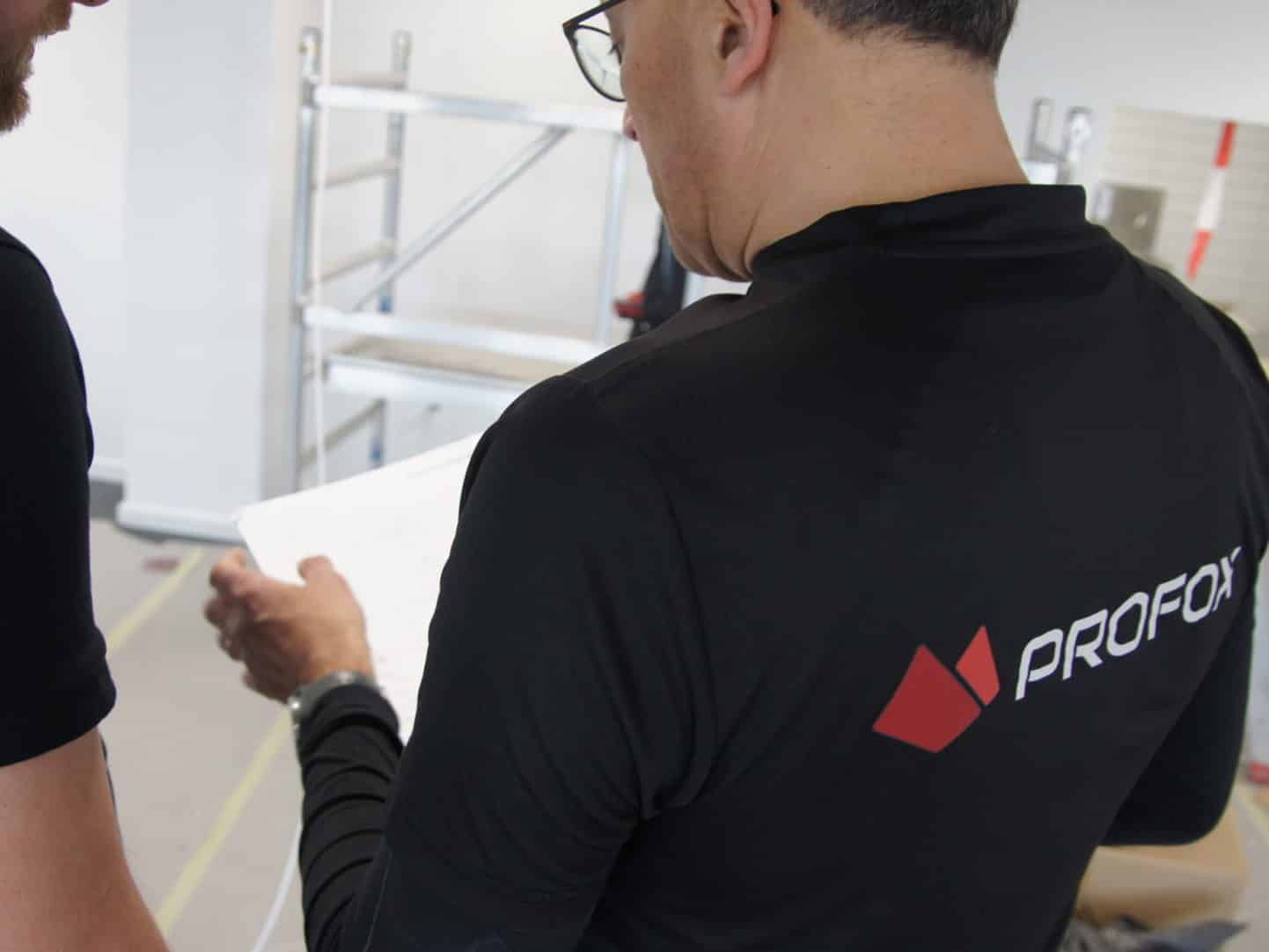 Two people stand indoors reviewing a document. One wears a black shirt with the PROFOX logo. A metal scaffold and other equipment are visible in the background.