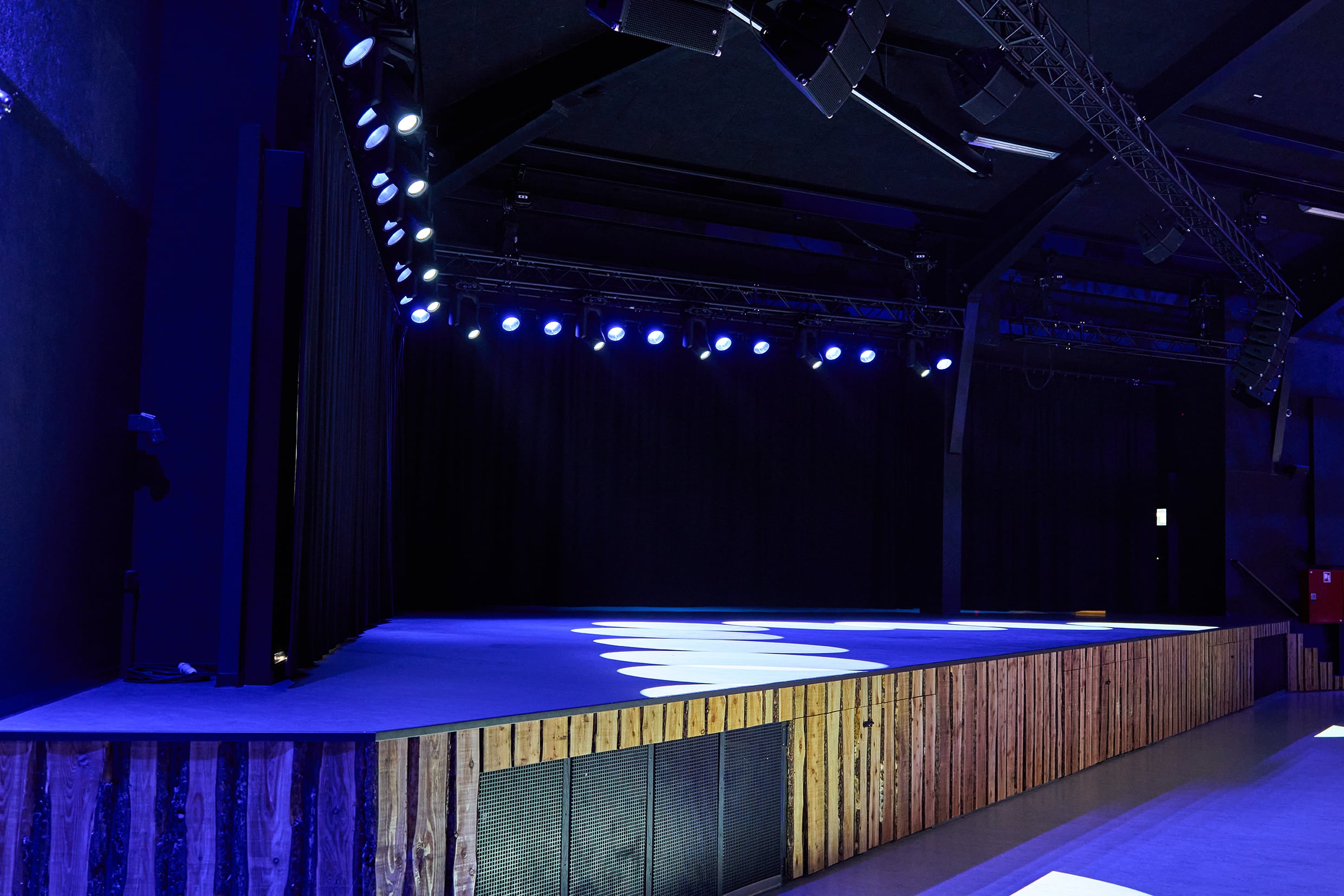 Empty stage with overhead spotlights shining down, creating bright patterns on the floor. The background is dark with curtains, and the sides of the stage are wooden. No people are present.