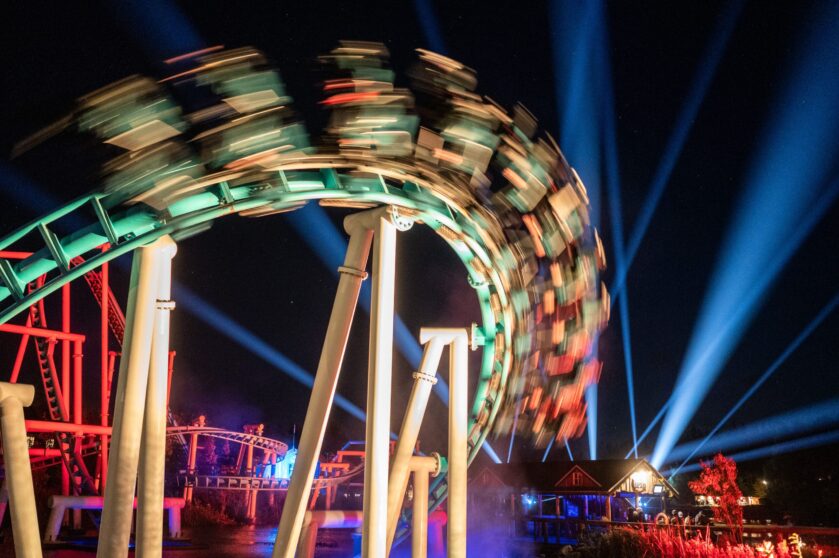 A roller coaster speeds along a curved track at night, lights creating motion blur. Blue beams shine into the sky and colorful lights illuminate the scene, with a building and trees in the background.