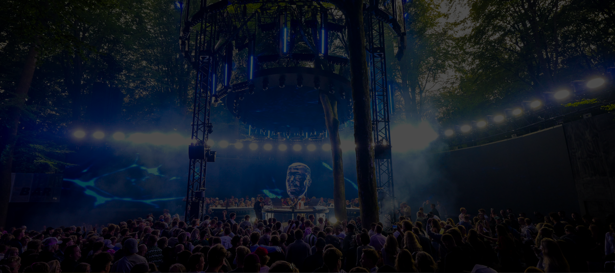 A large crowd faces a circular stage in a forest setting, featuring a DJ booth and a large EKKO SCREEN displaying a portrait.