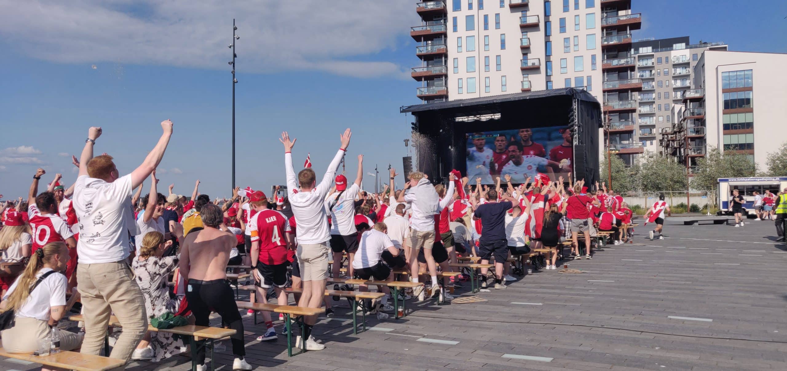 A crowd of people in red shirts cheer and raise their arms while watching a sports match on a large outdoor screen, with tall buildings in the background.