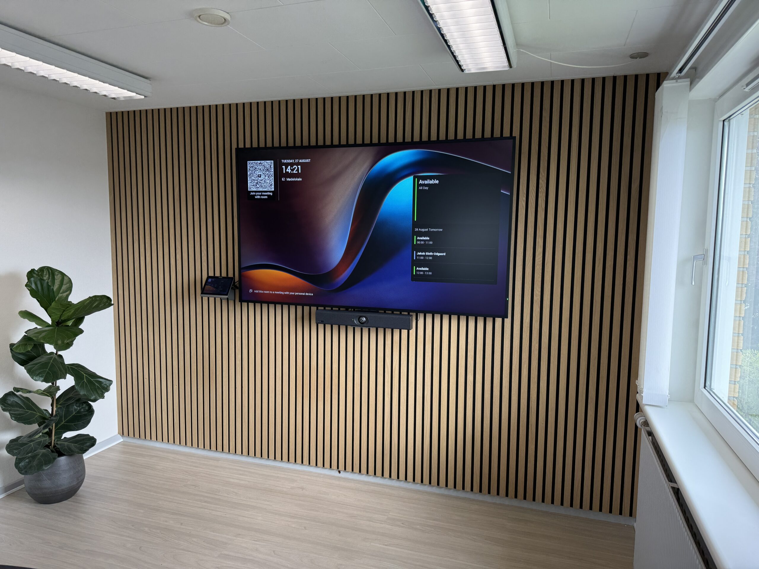Modern office corner with a wall-mounted flat screen displaying a conference interface, a soundbar below, wooden slat wall behind, potted plant on the left, and large windows on the right.