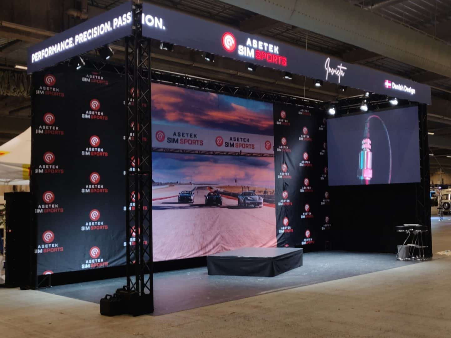 A trade show booth for Asetek SimSports features a stage, a large racing scene backdrop, branded banners, spotlights, and a screen displaying a close-up image of equipment. A small table and stools are on the right.