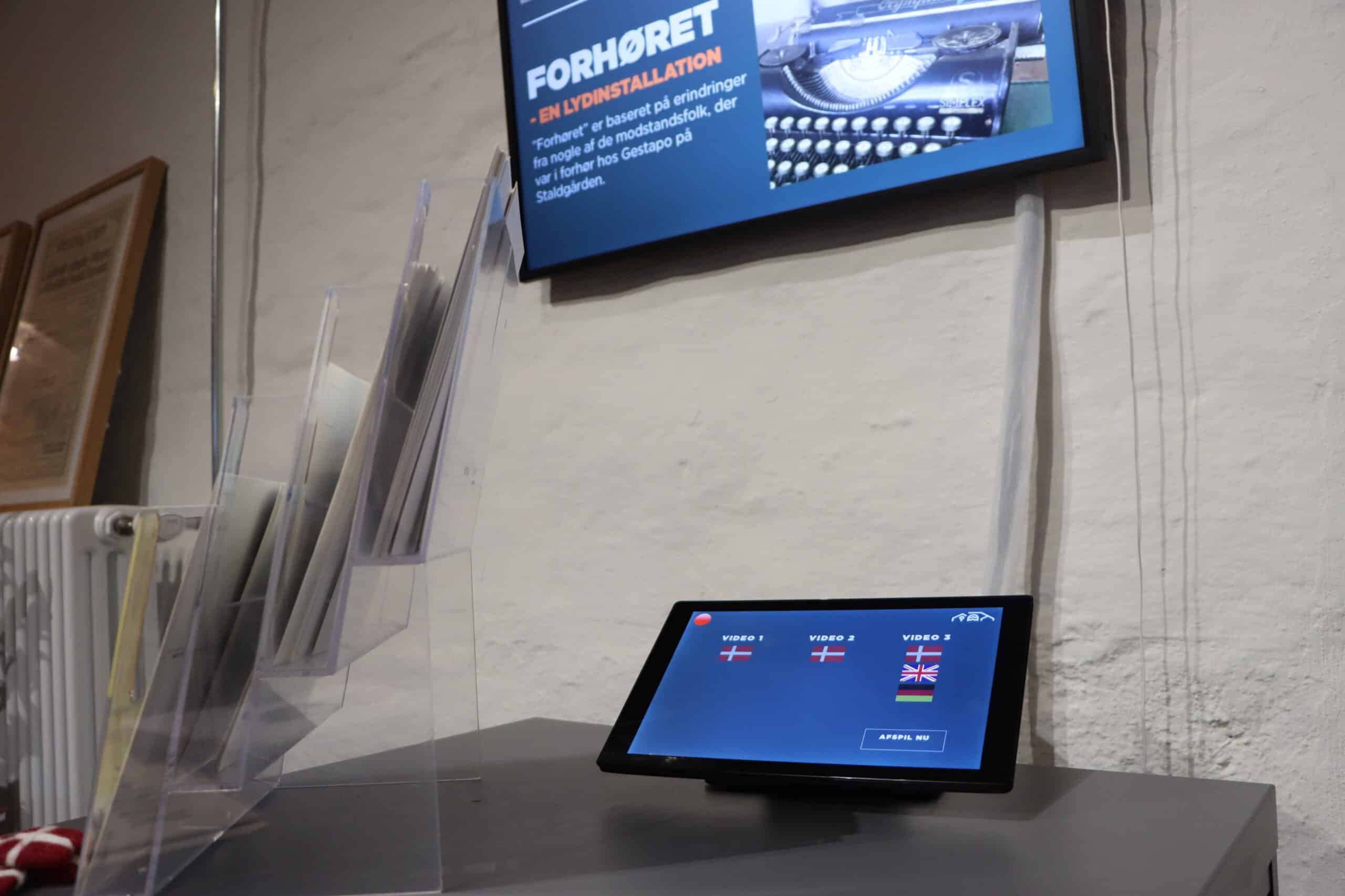 A touchscreen tablet on a stand displays language options with flags; behind it is a clear holder containing papers and a mounted screen with Danish text and an image of a typewriter.