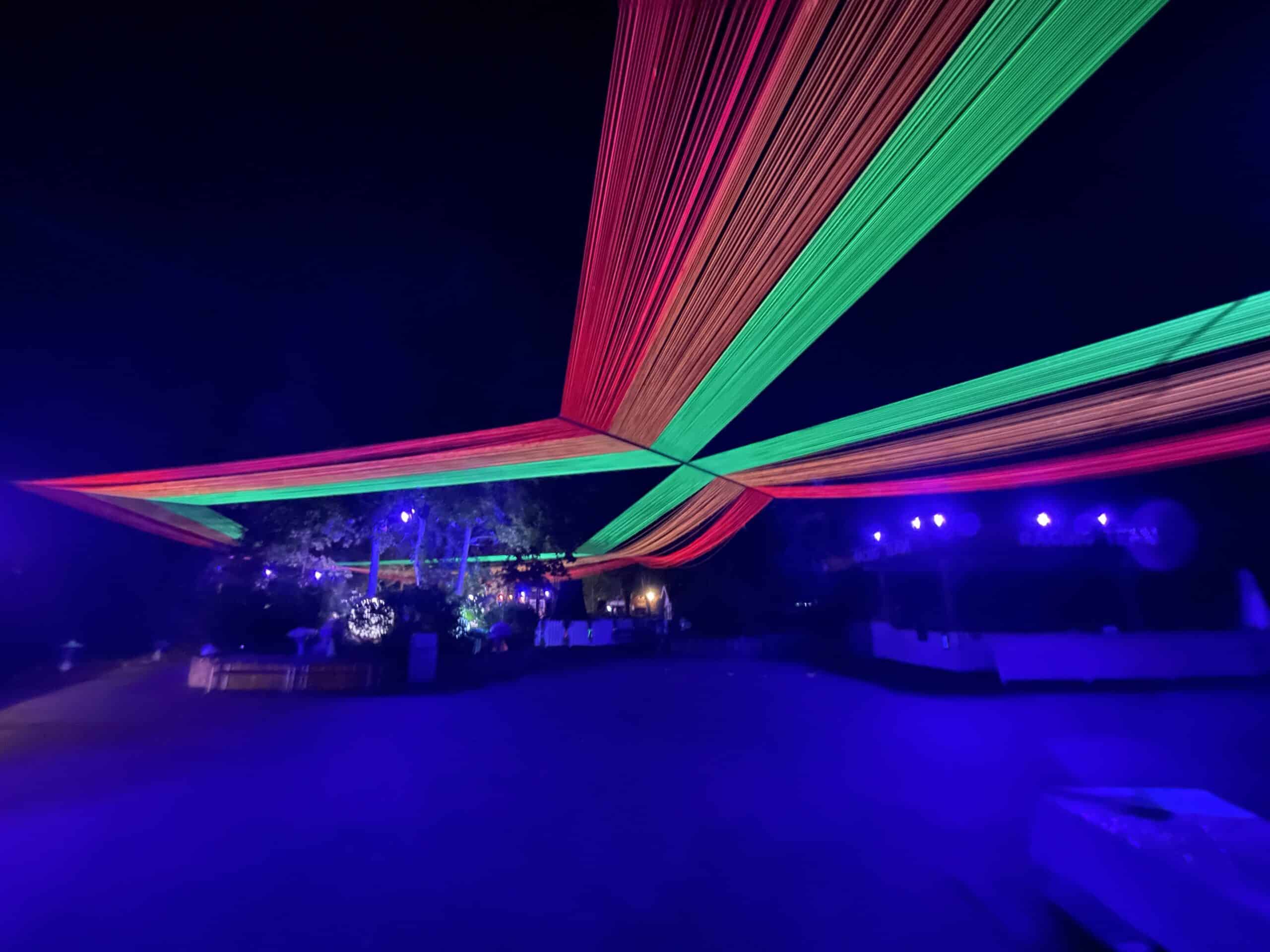 A nighttime scene with vibrant green and red fabric strands stretching overhead, illuminated by purple and blue lights, creating a festive atmosphere. Trees and seating are dimly visible in the background.