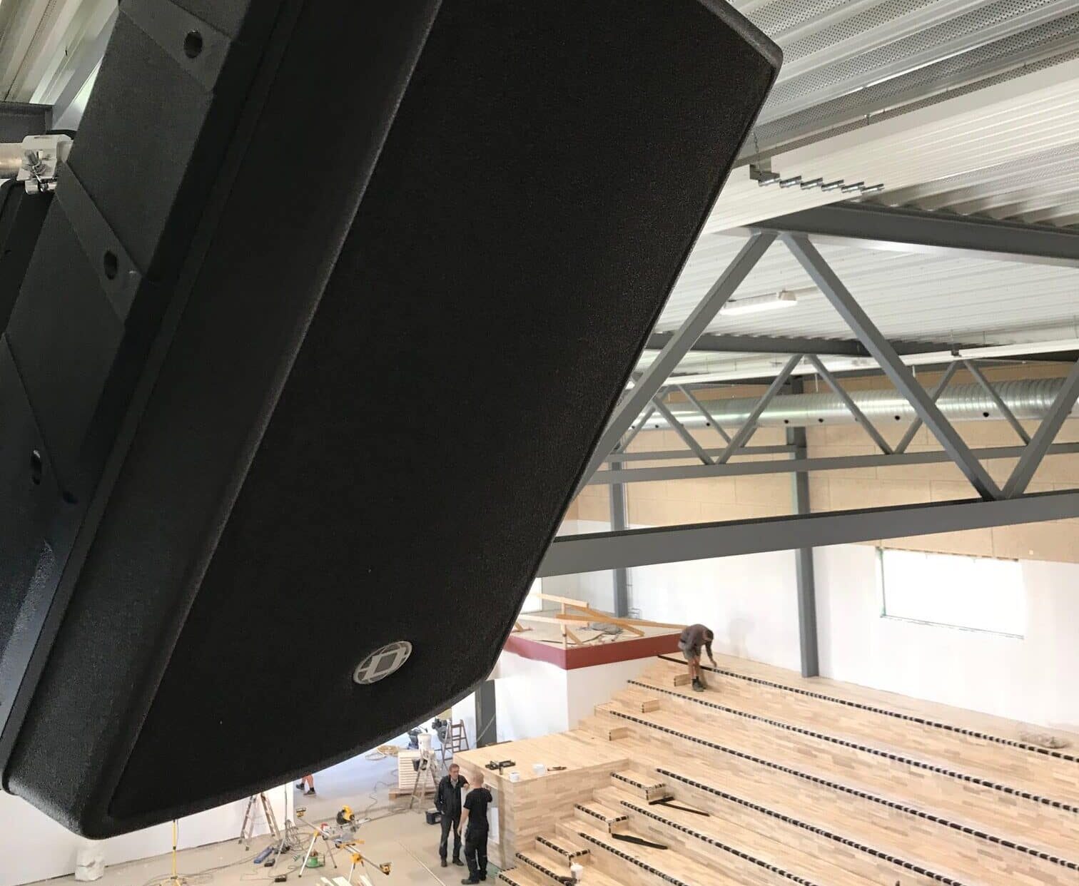A large black speaker is mounted in a metal-framed gym under construction, with workers on wooden bleachers and equipment scattered on the floor below.