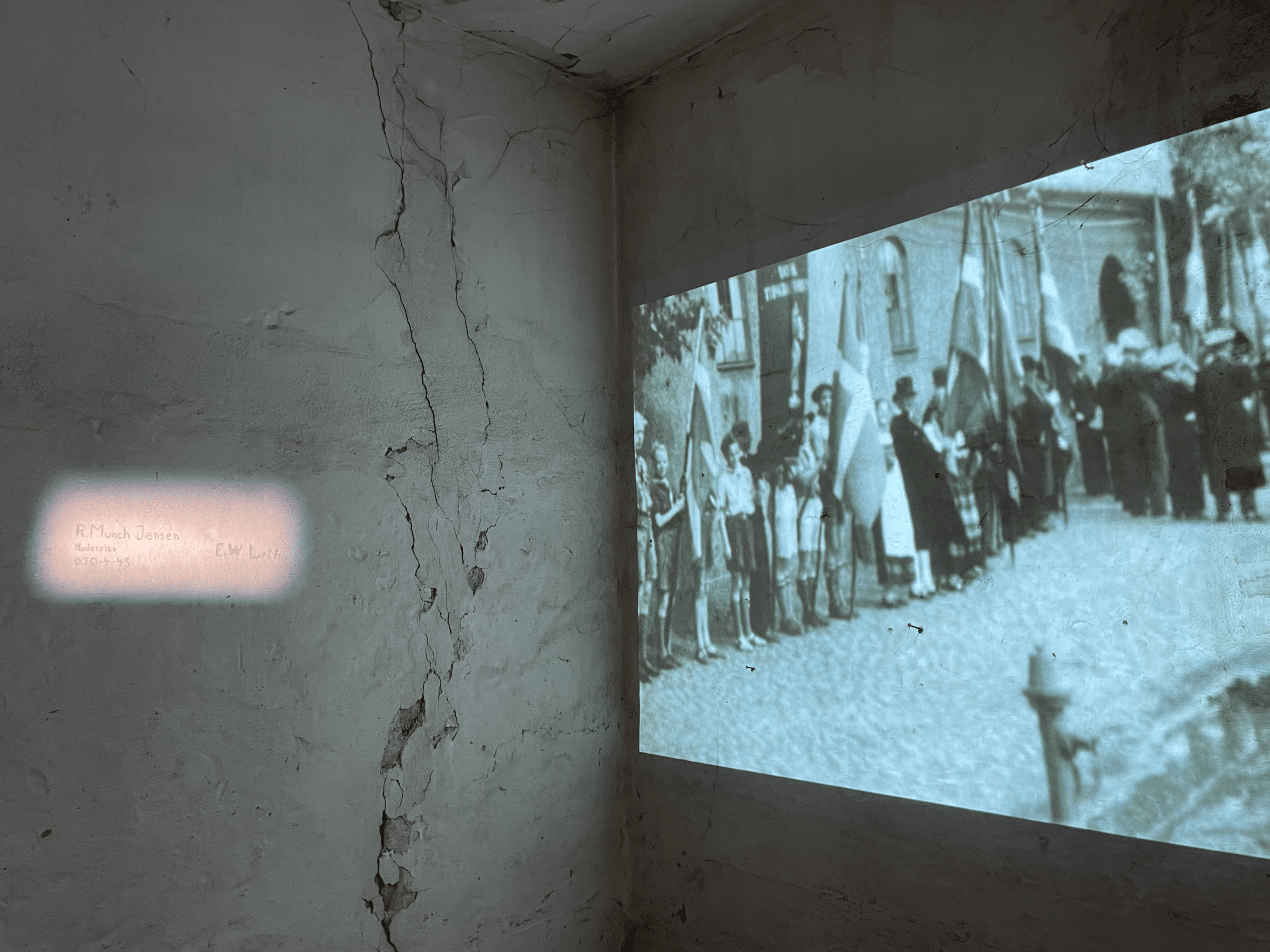 Black-and-white film is projected onto a cracked, worn wall, showing a group of people with flags in an outdoor scene; a small text projection appears on the left side of the wall.