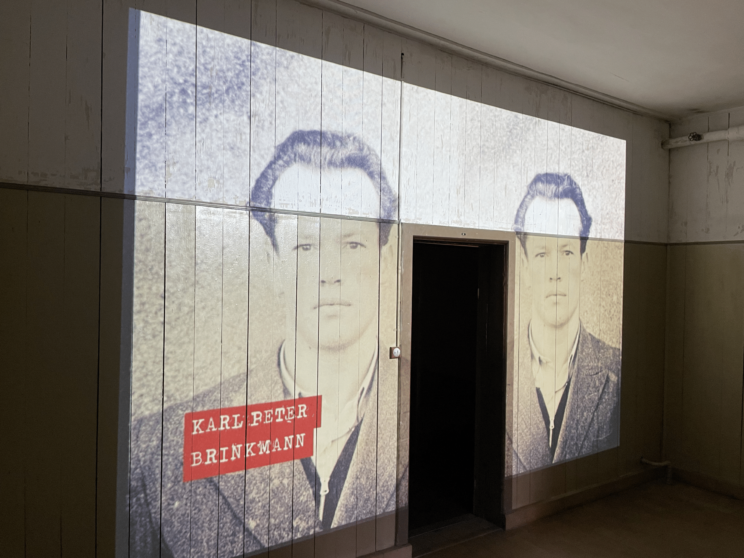 A black-and-white photo of a man is projected onto a wall above and beside a doorway, with the name Karl Peter Brinkmann in red text across the left image.